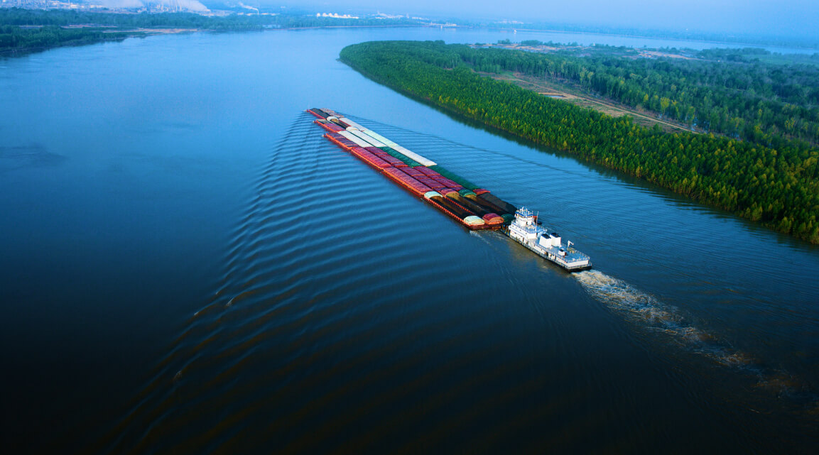 A barge transporting a large cargo of shipping containers on a wide stretch of the Mississippi river surrounded by lush forest